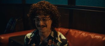 Movie still from “Weird: The Al Yankovic Story” (2022), directed by Eric Appel – A man with curly brown hair and glasses sitting on a red couch; Close Up shot, High angle