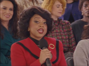 Movie still from “Weird: The Al Yankovic Story” (2022), directed by Eric Appel – A woman in a red jacket is holding a microphone; Medium shot, Over the shoulder angle