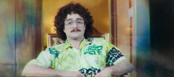 Movie still from “Weird: The Al Yankovic Story” (2022), directed by Eric Appel – A man sitting in a chair wearing a hawaiian shirt; Medium shot, High angle