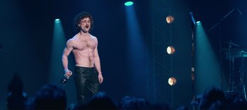 Movie still from “Weird: The Al Yankovic Story” (2022), directed by Eric Appel – A man with curly red hair and no shirt is on a stage; Wide shot, Over the shoulder angle