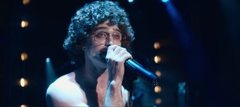 Movie still from “Weird: The Al Yankovic Story” (2022), directed by Eric Appel – A man with curly hair is singing into a microphone; Medium shot, Low angle