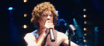 Movie still from “Weird: The Al Yankovic Story” (2022), directed by Eric Appel – A man with curly hair and glasses is singing into a microphone; Medium shot, Low angle