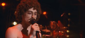 Movie still from “Weird: The Al Yankovic Story” (2022), directed by Eric Appel – A man with curly hair and glasses is holding a microphone; Close Up shot, Over the shoulder angle