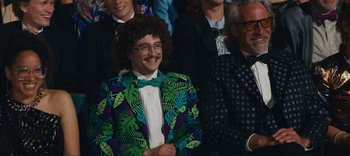 Movie still from “Weird: The Al Yankovic Story” (2022), directed by Eric Appel – A man in a suit and bow tie is sitting in front of a group of people; Medium shot, Over the shoulder angle