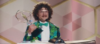 Movie still from “Weird: The Al Yankovic Story” (2022), directed by Eric Appel – A man with a curly wig and a green bow tie holding a bottle of champagne; Medium shot, Low angle
