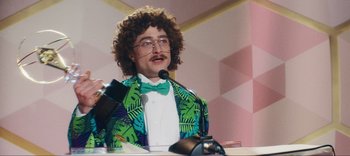 Movie still from “Weird: The Al Yankovic Story” (2022), directed by Eric Appel – A man with a curly hair and a green bow tie is giving a speech; Medium shot, Low angle