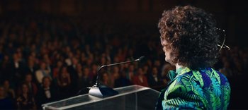 Movie still from “Weird: The Al Yankovic Story” (2022), directed by Eric Appel – A woman is giving a speech to a crowd of onlookers; Medium shot, Over the shoulder angle