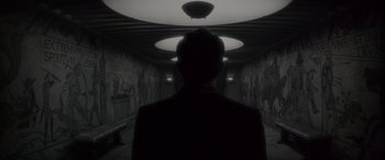 Movie still from “Werewolf by Night” (2022), directed by Michael Giacchino – A man standing in a dark hallway with lights above him; Wide shot, Low angle