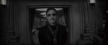 Movie still from “Werewolf by Night” (2022), directed by Michael Giacchino – A man in a black suit with a necklace in a room; Medium shot, Over the shoulder angle