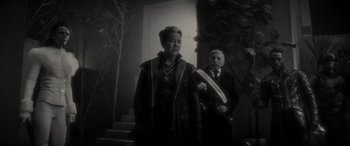 Movie still from “Werewolf by Night” (2022), directed by Michael Giacchino – An older woman standing next to an older man; Medium shot, Over the shoulder angle