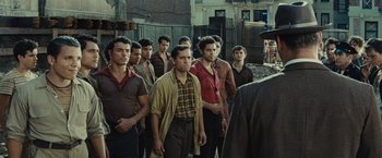 Movie still from “West Side Story” (2021), directed by Steven Spielberg – A group of men standing next to each other in front of a building; Medium shot, Over the shoulder angle