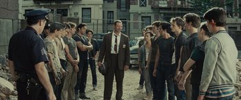 Movie still from “West Side Story” (2021), directed by Steven Spielberg – A group of young men standing around a man in a brown suit; Wide shot, Over the shoulder angle