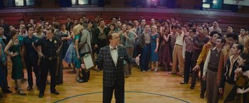 Movie still from “West Side Story” (2021), directed by Steven Spielberg – A crowd of people standing in a room with a man in a plaid suit; Wide shot, High angle