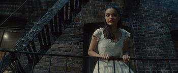 Movie still from “West Side Story” (2021), directed by Steven Spielberg – A woman in a white dress standing next to a staircase; Medium shot, Low angle