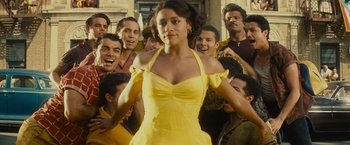 Movie still from “West Side Story” (2021), directed by Steven Spielberg – A woman in a yellow dress standing in front of a group of men; Medium shot, Over the shoulder angle