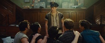Movie still from “West Side Story” (2021), directed by Steven Spielberg – A group of young men sitting in front of a man in a hat; Medium shot, Over the shoulder angle