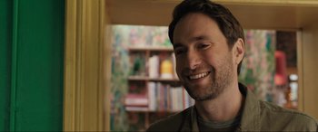 Movie still from “What's Love Got to Do with It?” (2022), directed by Shekhar Kapur – A man smiling for the camera in front of bookshelves; Close Up shot, Over the shoulder angle