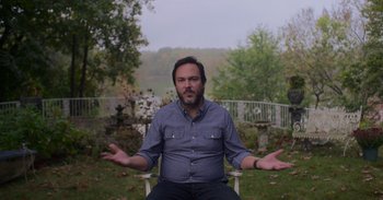 Movie still from “White Hot: The Rise & Fall of Abercrombie & Fitch” (2022), directed by Alison Klayman – A man sitting in a chair in the grass shrubbery; Medium shot, High angle