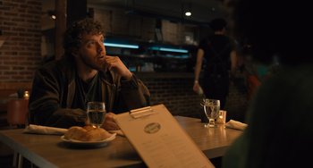 Movie still from “White Men Can't Jump” (2023), directed by Calmatic – A man sitting at a table in front of a plate of food; Medium shot, Over the shoulder angle