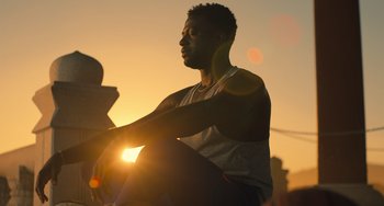 Movie still from “White Men Can't Jump” (2023), directed by Calmatic – A man sitting on top of a bench at sunset; Medium shot, Low angle