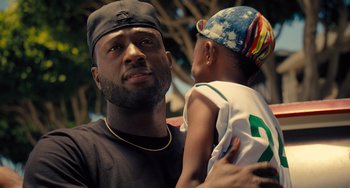 Movie still from “White Men Can't Jump” (2023), directed by Calmatic – A man holding a child in his arms and smiling; Close Up shot, Over the shoulder angle