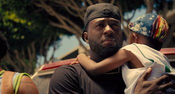 Movie still from “White Men Can't Jump” (2023), directed by Calmatic – A man holding a child in his arms and wearing a baseball cap; Close Up shot, Over the shoulder angle