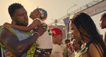 Movie still from “White Men Can't Jump” (2023), directed by Calmatic – A man holding a child in his arms while a crowd looks on; Medium shot, Over the shoulder angle