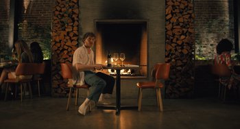 Movie still from “White Men Can't Jump” (2023), directed by Calmatic – A man sitting in front of a fireplace with a glass of wine in front of him; Wide shot, Low angle