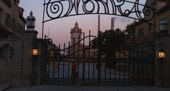 Movie still from “Willy Wonka & the Chocolate Factory” (1971), directed by Mel Stuart – A person walking through a gate with a clock tower in the background; Extreme Wide shot, Low angle