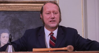 Movie still from “Willy Wonka & the Chocolate Factory” (1971), directed by Mel Stuart – A man in a suit and tie sitting in front of a microphone; Close Up shot, Low angle