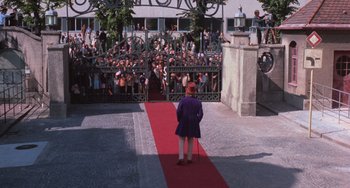 Movie still from “Willy Wonka & the Chocolate Factory” (1971), directed by Mel Stuart – A man in a purple coat and a red hat is standing on a red carpet in front of a crowd of onlookers; Wide shot, High angle