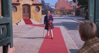 Movie still from “Willy Wonka & the Chocolate Factory” (1971), directed by Mel Stuart – A man in a purple coat and a cane standing on a red carpet in front of a building; Wide shot, High angle