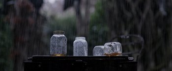 Movie still from “Winnie the Pooh: Blood and Honey” (2023), directed by Rhys Frake-Waterfield – Three glass jars sitting on top of a wooden table in the rain; Extreme Close Up shot, Low angle