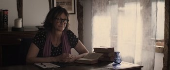 Movie still from “Winnie the Pooh: Blood and Honey” (2023), directed by Rhys Frake-Waterfield – A woman sitting at a table in front of a stack of books; Medium shot, Low angle