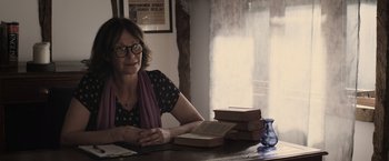 Movie still from “Winnie the Pooh: Blood and Honey” (2023), directed by Rhys Frake-Waterfield – An older woman sitting at a table in front of a stack of books; Medium shot, Over the shoulder angle