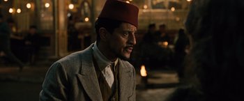 Movie still from “Wonder Woman” (2017), directed by Patty Jenkins – A man wearing a fez and a suit in a dimly - lit room; Close Up shot, Over the shoulder angle