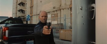 Movie still from “Wrath of Man” (2021), directed by Guy Ritchie – A man holding a gun while standing in front of a building; Medium shot, Low angle