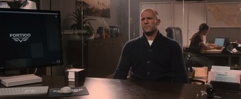 Movie still from “Wrath of Man” (2021), directed by Guy Ritchie – A bald man sitting at a table in a room; Medium shot, Over the shoulder angle