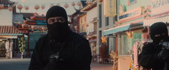 Movie still from “Wrath of Man” (2021), directed by Guy Ritchie – A man wearing a black mask and holding a camera in front of buildings; Medium shot, Low angle