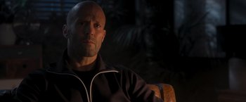 Movie still from “Wrath of Man” (2021), directed by Guy Ritchie – A bald man sitting in front of a palm tree; Close Up shot, Over the shoulder angle