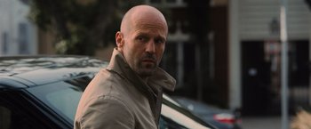 Movie still from “Wrath of Man” (2021), directed by Guy Ritchie – A man with a bald head standing in front of a car; Close Up shot, Over the shoulder angle