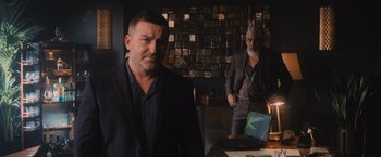 Movie still from “Wrath of Man” (2021), directed by Guy Ritchie – Two men standing in front of a laptop computer; Medium shot, Over the shoulder angle