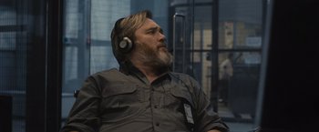 Movie still from “Wrath of Man” (2021), directed by Guy Ritchie – A man with a beard wearing headphones sitting in a room; Close Up shot, Over the shoulder angle