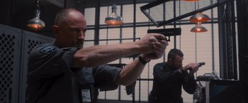 Movie still from “Wrath of Man” (2021), directed by Guy Ritchie – A man holding a gun in a room with two other men; Medium shot, Low angle