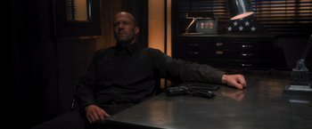 Movie still from “Wrath of Man” (2021), directed by Guy Ritchie – A man sitting at a table holding a gun in his hand; Medium shot, Over the shoulder angle