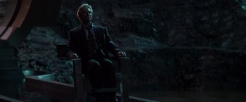 Movie still from “X-Men” (2000), directed by Bryan Singer – A man in a suit and tie sitting in a chair in front of a rock wall; Wide shot, High angle