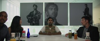 Movie still from “Yesterday” (2019), directed by Danny Boyle – A man sitting at a table in front of a wall with a picture of himself; Medium shot, Over the shoulder angle