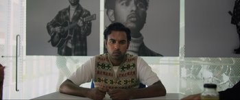 Movie still from “Yesterday” (2019), directed by Danny Boyle – A man sitting at a table in front of a poster; Medium shot, Over the shoulder angle