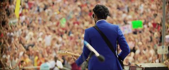 Movie still from “Yesterday” (2019), directed by Danny Boyle – A man in a blue suit is playing a guitar in front of a large crowd; Medium shot, Over the shoulder angle
