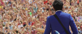 Movie still from “Yesterday” (2019), directed by Danny Boyle – A man in a blue suit is playing a guitar in front of a large crowd; Medium shot, Over the shoulder angle